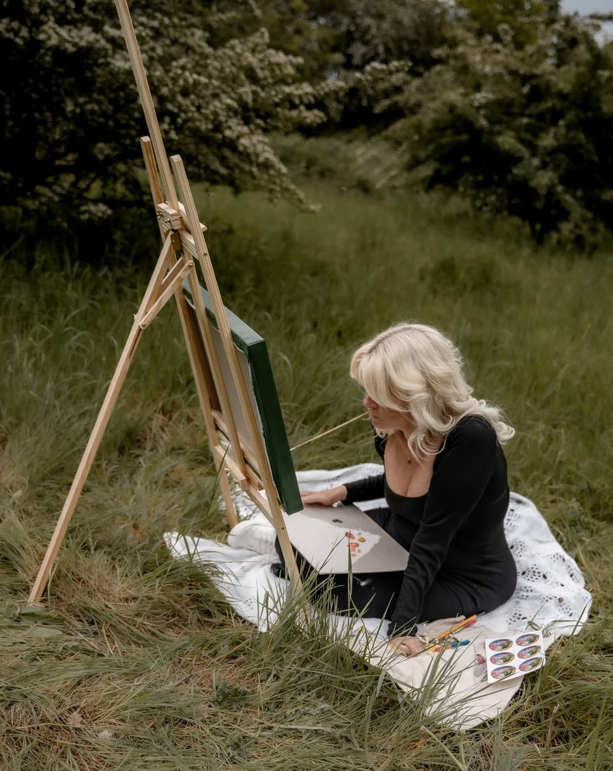 Elīna Veinberga painting at an easel outdoors in a meadow